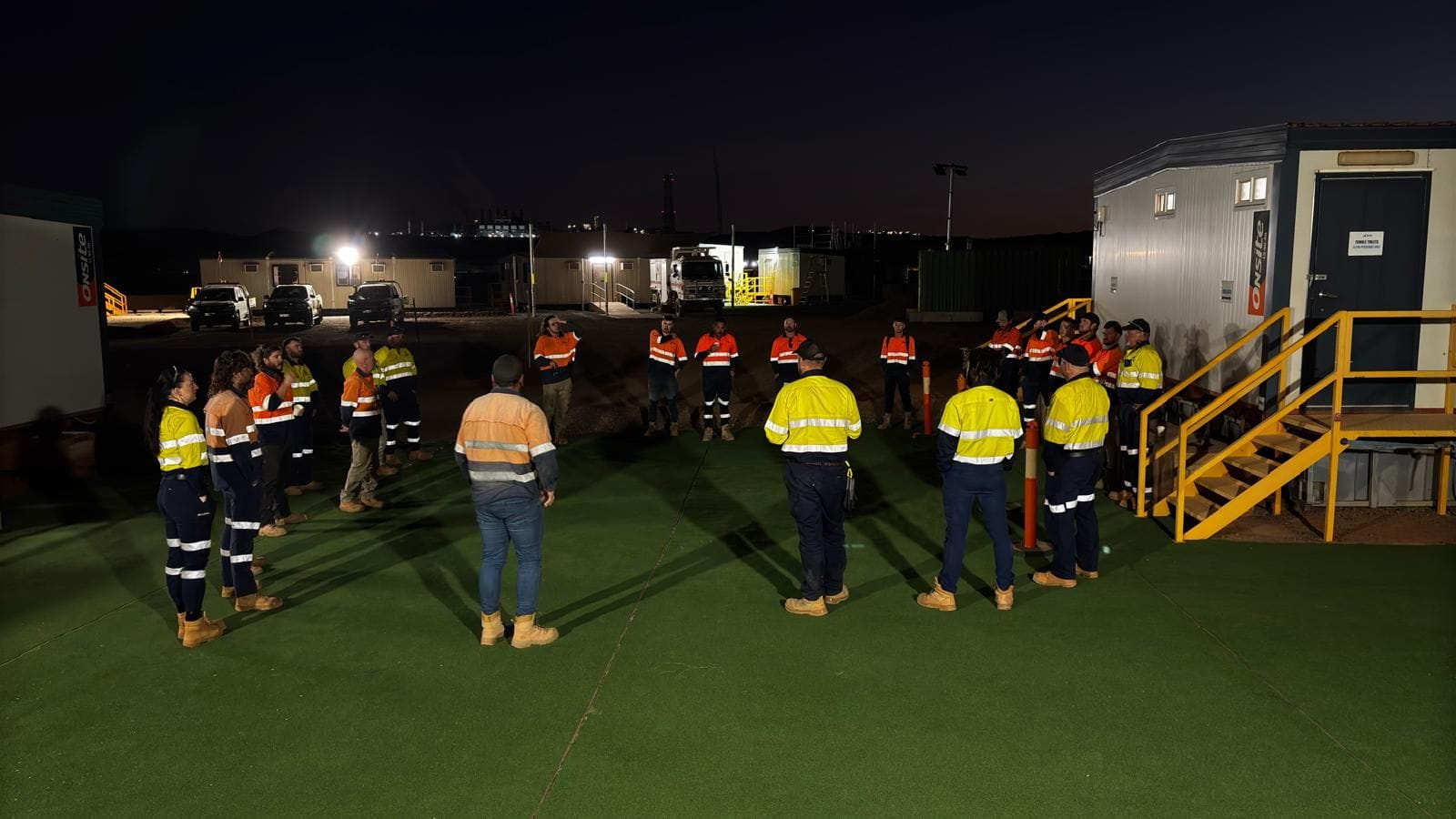 Crew gathered for a night pre-start briefing outside site offices with plant lit on the horizon